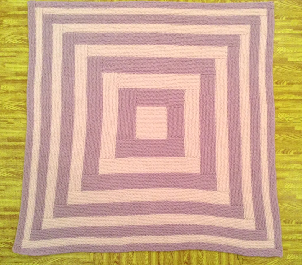 A knitted blanket with concentric squares in alternating light pink and lavender colors placed on a wooden floor.