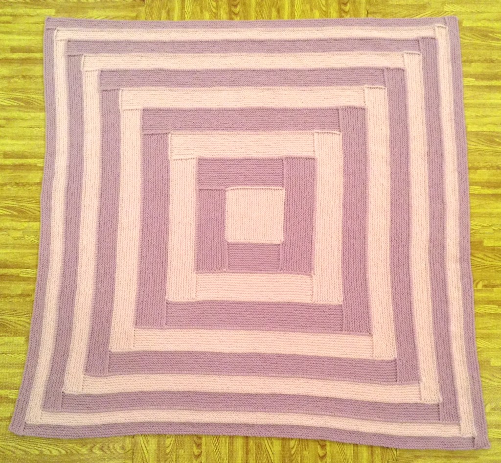 A knitted blanket with a geometric pattern featuring concentric squares in shades of pink and lavender, placed on a wooden floor.